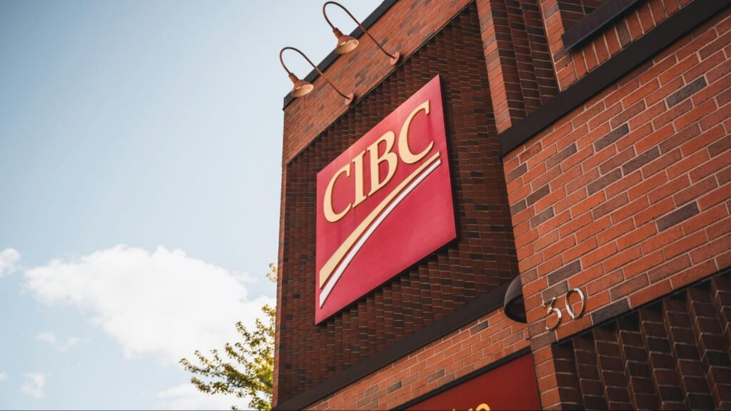 Close up of CIBC logo mounted on a brick bank building exterior beneath overhead lamps and blue sky backdrop